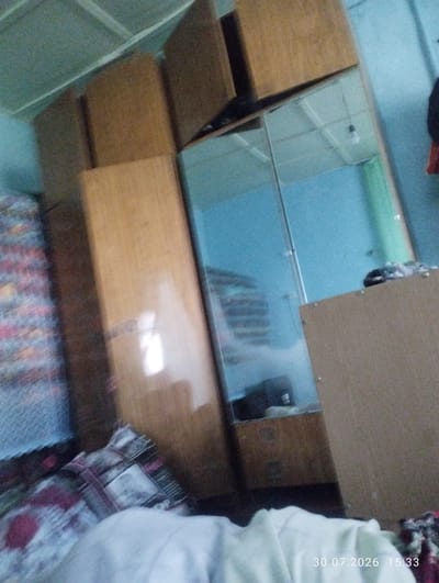 The image is a low-quality, blurry photograph taken indoors, likely in a residential setting in Quyosh, Uzbekistan. The scene appears to be a bedroom, with furniture and bedding visible.
Dominating the center of the frame is a large wooden wardrobe with mirrored doors. The wardrobe is tall and appears to have multiple sections. One section of the wardrobe is open, revealing the interior, which contains shelves and possibly some stored items. The mirror reflects the ceiling, which has a light fixture, and a light blue wall.
To the left of the wardrobe, there is a mattress or bed with patterned bedding. The pattern is indistinct due to the blurriness.
To the right of the wardrobe, there is another piece of wooden furniture, possibly a dresser or another part of the wardrobe unit. This furniture has a flat, light brown surface.
The overall environment appears somewhat cluttered, with a general sense of disarray. The lighting is dim, contributing to the blurriness of the image. There are no people clearly visible in the photograph. The date stamp "30.07.2026 15:33" is superimposed on the lower right corner of the image.