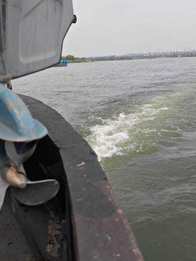 The image shows a view from the stern of a boat on a river, with a wake trailing behind. In the foreground, the dark railing of the boat is visible, along with a portion of a blue mechanical component, possibly part of the engine or propeller assembly. The water is a murky green color, and the wake is turbulent with white foam. In the background, across the river, is a tree-lined shore with a large industrial complex on the horizon, characterized by numerous smokestacks. A small blue boat is visible in the distance to the left. The sky is overcast. This scene is consistent with the industrial areas along the river near Peschanaka, Russia, which is known for its heavy industry and proximity to waterways.