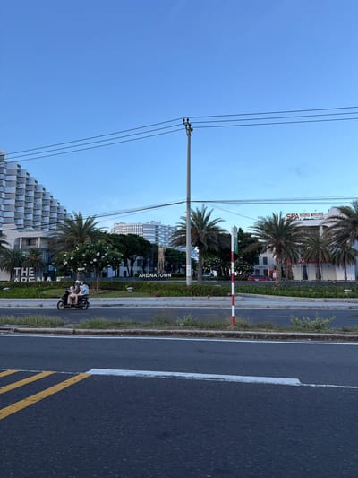 In Cam Ranh Ward, Vietnam, a street scene unfolds under a clear blue sky. On the left, a modern, multi-story building with a distinctive geometric facade dominates the background. In front of it, a sign reads "THE ARENA". To the right of this building, a road runs horizontally across the image. On this road, a motorbike with three occupants is traveling from left to right. The occupants are wearing casual clothing and helmets.
Further to the right, another building is visible with a sign that says "SPA HỒNG NHUNG LUXURY". Palm trees are scattered throughout the scene, adding to the tropical ambiance. A tall utility pole with multiple wires stretches across the sky. In the foreground, the road surface is visible, marked with yellow and white lines. A red and white striped pole stands on the side of the road. Greenery and small bushes line the median of the road and the area behind it. The lighting suggests it is daytime, possibly late afternoon.