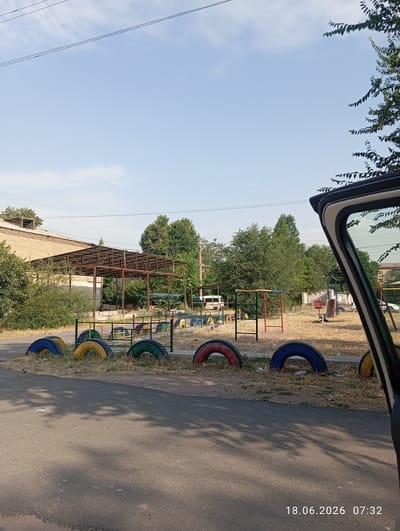 The image depicts an outdoor scene in the city of Ohangaron, Uzbekistan, on June 18, 2026, at 07:32. In the foreground, a paved asphalt surface with shadows cast by unseen trees is visible. Along the edge of the asphalt, several colorful tires, painted blue, yellow, and red, are embedded in the dry, grassy ground, serving as a decorative border.

Beyond the tires, a children's playground is situated on a patch of dry grass and dirt. Various playground equipment can be identified, including a red and green climbing frame, a blue swing set, and what appears to be a see-saw or a similar apparatus. A small white van is parked in the background to the left, near a brick building with a corrugated metal roof structure extending from it. Tall green trees are scattered throughout the area, and a clear blue sky with a few faint clouds is overhead. The overall impression is that of an outdoor recreational space in a somewhat arid environment. The perspective is from within a vehicle, as indicated by the car door frame visible on the right side of the image.