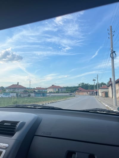 The image depicts a view from inside a vehicle traveling through a village. The road ahead is paved and curves slightly to the right. On the left side of the road, there are houses with red-tiled roofs and a well-maintained fence. A silver car is parked on the left. On the right side of the road, there are more houses, some appearing older and in disrepair, with visible power lines and a utility pole. A stork's nest is visible atop a utility pole on the right. The sky is blue with scattered white clouds, some appearing wispy and elongated. The foreground shows the interior of the car's dashboard, including an air vent and buttons. The location is identified as Novo Zhelezare, Bulgaria.