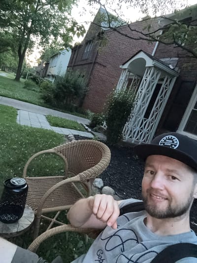 A Caucasian man with short brown hair, a beard, and brown eyes is sitting in a wicker chair in a yard. He is wearing a grey t-shirt with a black design and a black baseball cap with a white logo. He is smiling at the camera. In the background, there is a brick house with a white porch and trees. The setting appears to be a residential neighborhood in Shaker Heights, Ohio.