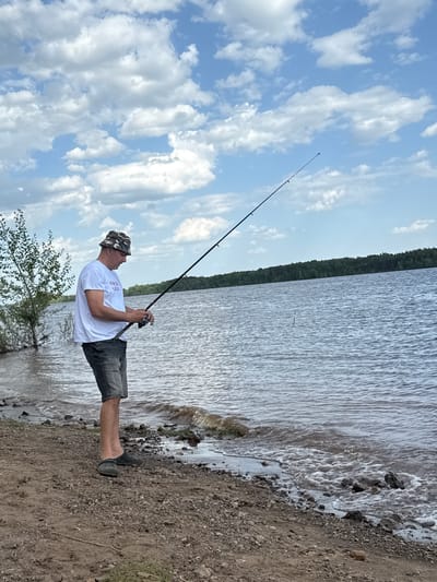 A man is fishing at Doksha Lake in Doksha, Russia. The man is wearing a white t-shirt, grey shorts, a camouflage bucket hat, and black shoes. He is holding a fishing rod. The lake is in the background, with a shoreline covered in pebbles. The sky is blue with white clouds.