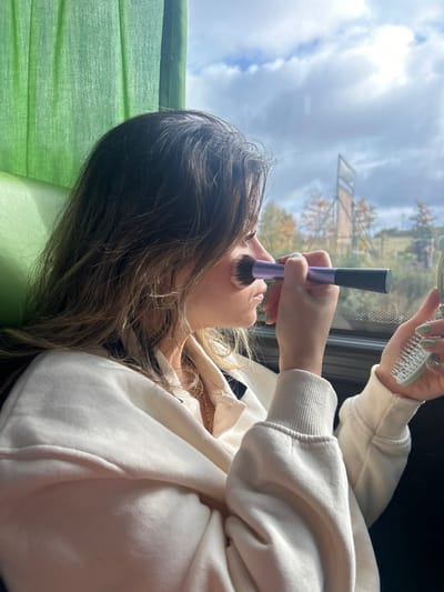 A young female, viewed in profile from the right, is seated inside a vehicle, identified as being in Kaplice, Czechia. She has long, light brown hair with blonde highlights and wears a cream-colored collared sweatshirt, with a thin gold chain visible around her neck. Her left hand holds a purple-handled makeup brush with dark bristles, which is positioned against her right cheek. Her right hand, displaying light green nail polish, holds a small, light green textured object, possibly a compact mirror or a small hairbrush. The interior of the vehicle shows a green fabric curtain and a green upholstered seat on the left. Through the window, a daylight scene is visible with a blue sky partially obscured by white clouds, autumnal trees displaying yellow and orange foliage, and a prominent dark rectangular structure in the mid-ground, against a backdrop of distant hills.