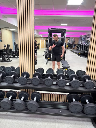 The image captures an indoor fitness gym located in Babergh, United Kingdom. A male subject, appearing to be in his 30s or 40s, with short dark hair, glasses, and a short beard, is depicted in a mirror selfie. He is wearing a black short-sleeved Nike t-shirt, black shorts, and a silver chain necklace. He holds a smartphone in his right hand to capture his reflection, with his left hand resting on his hip.
In the immediate foreground, a two-tiered black metal rack displays numerous black Technogym dumbbells with white weight markings. The visible dumbbells on the top tier include 5kg, 6kg, 7kg, 8kg, 9kg, and 10kg, while the bottom tier shows 20kg, 22kg, 24kg, 26kg, and 28kg.
The mirror's reflection reveals a modern gym environment. Behind the primary subject, a black power rack equipped with a barbell and weight plates stands alongside a grey adjustable weight bench. To the right, a row of treadmills and other cardio machines extends into the background. To the far left in the reflection, another male individual, wearing a black cap and dark clothing, is partially visible near additional weight equipment, appearing to handle a barbell.
The gym features a light grey speckled floor, light-colored walls, some with light blue sections, and striking pink LED strip lighting along the ceiling edges, interspersed with white square panel lights. A light wooden door marked with an "EXIT" sign is visible in the background reflection.