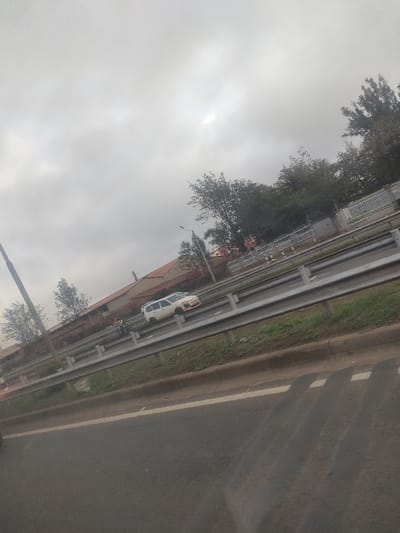 This image depicts a multi-lane road environment in Gatongora ward, Kenya, under an overcast sky. In the mid-ground, a white compact SUV, resembling a Toyota Probox, is positioned on an elevated section of a highway. Two individuals are visible near the driver's side of this white SUV; one is distinctly wearing a yellow/green high-visibility vest. The elevated roadway is separated by a metal guardrail from the lower road in the foreground. Beyond the elevated road, several trees and structures, including buildings with brown roofs and a white fence, are visible. A streetlight pole stands on the left. The foreground shows an asphalt road surface with a solid white line and tire marks/shadows.