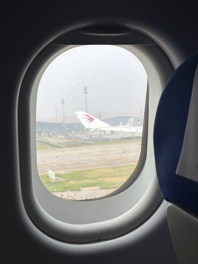 The image shows a view from an airplane window, looking out at an airport tarmac. The window has condensation and water droplets on it. In the background, a white airplane with a red and blue logo on its tail is visible. Other airport structures, such as buildings and light poles, can also be seen. The weather appears overcast. On the right side of the image, a portion of an airplane seat with blue upholstery is visible. The scene is likely at Chengdu Shuangliu International Airport in Guangyang District, China, given the presence of airplanes and airport infrastructure.