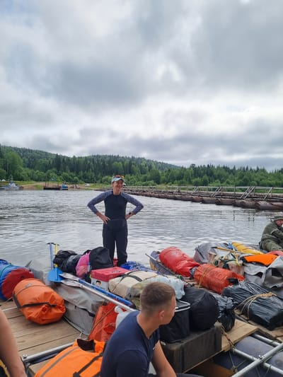 A group of people are on a raft loaded with gear on a river. A woman in a dark blue long-sleeved shirt with a white geometric pattern and black pants stands in the center of the raft, facing to the right with her hands on her hips. A man in a green jacket sits on the raft to the right, looking towards the water. Several people are partially visible on the left and right sides of the raft. The raft is filled with various bags, containers, and equipment, including orange dry bags, black duffel bags, a pink backpack, a red box, and a blue paddle. In the background, a dense green forest lines the riverbank. A long structure, appearing to be a pontoon bridge made of logs and barrels, extends across the river. Several boats are docked on the left bank. The sky is overcast with clouds. The location is the Visherogorsk, Russia.