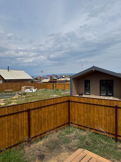 This is a landscape photograph taken in Khuzhir, Russia, depicting a residential area with a focus on fences and buildings under a cloudy sky. In the foreground, a tall, wooden fence with a dark metal frame runs diagonally across the frame, creating a sense of depth. Beyond this fence, another wooden fence encloses a yard with dry grass and some green patches.
To the left, a single-story wooden house with a white, corrugated metal roof and a visible chimney is situated. Adjacent to it is a wooden bench placed near a small body of water, possibly a pond. Several white sacks and wooden beams are also present in this area, suggesting ongoing construction or material storage.
In the middle ground, a cluster of houses with colorful roofs (red, blue, and green) are visible, indicating a village or small town setting. In the right portion of the frame, a modern, two-story house with a dark brown exterior and a dark gray roof stands out. This house features large windows and a wooden deck. A small black cat is perched on the deck railing of this modern house, facing left. Further in the background, a tall, slender tower, possibly a communication tower, is visible against the sky. The horizon shows rolling hills and a body of water, likely Lake Baikal, given the location. The sky is predominantly covered with light gray clouds, with some patches of blue peeking through.