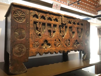 This image shows an ornate, antique wooden chest sitting on a shelf. The chest is dark brown, likely from age and polishing, and features numerous carved details. On the sides are circular patterns and the front has a series of shield-shaped carvings, with additional floral or geometric decorations between and beneath the shields. The chest appears to be in a museum or exhibit, as indicated by the "Do Not Touch" sign visible above it, and a modern black metal support structure can be seen in the background. The background appears to be an interior setting with bright, diffuse lighting. Considering the location in Veytaux, Switzerland, the chest is most likely on display at the Château de Chillon museum.