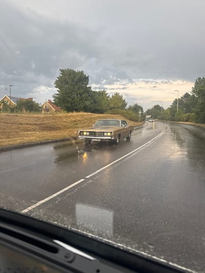 The image shows a gold-colored vintage American car driving on a wet road in Falkenberg, Sweden. The sky is overcast with dark clouds. The road is slick with rain, and reflections of streetlights and the car's headlights are visible on the surface. The car appears to be a Ford Galaxie from the late 1960s, identified by its distinctive grille and headlights. The license plate, partially visible, is B XL 268H.
On the left side of the road, there is a grassy embankment with some houses and trees in the background. On the right side of the road, there are more trees and streetlights. Another car is visible in the distance, also driving on the wet road. The perspective suggests the photo was taken from inside another vehicle. The overall scene depicts a gloomy, wet day in a suburban or semi-rural area of Falkenberg.