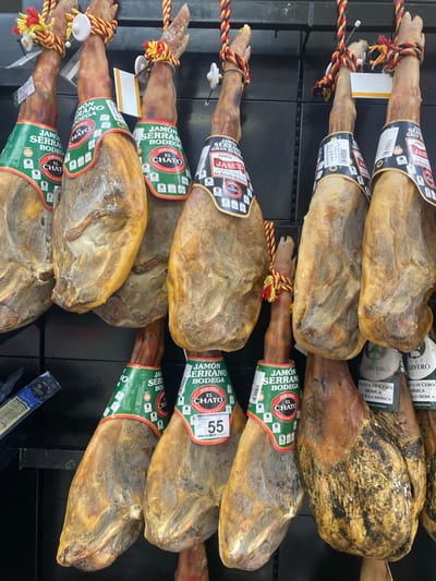 A supermarket display in Bel Air, Spain, features a row of cured hams, known as jamón serrano, hanging from hooks. Each ham is partially wrapped in green and red paper bands with Spanish text and branding. Some hams have yellow and red braided ropes attached to their tops. The background consists of a dark, tiled wall and shelving. The lighting is bright and appears to be overhead.