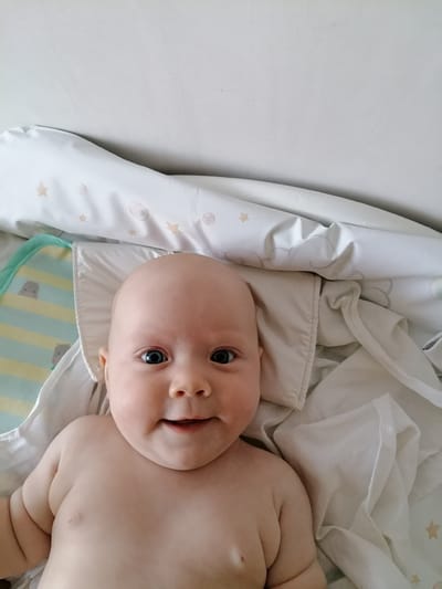 This is a close-up, high-angle shot of a baby lying on a bed. The baby, who appears to be a few months old, is bald with bright blue eyes and a happy smile. They are lying on their back, with their bare chest and shoulders visible. The baby is propped up on a pillow, and there is a blanket with a cloud and star pattern behind them. A striped blanket with cartoon hippos is partially visible on the left side of the frame. The background is a plain, light-colored wall, suggesting the image was taken indoors. The lighting appears to be natural, possibly from a window, and the overall mood is cheerful and intimate. There is no visible text in the image.