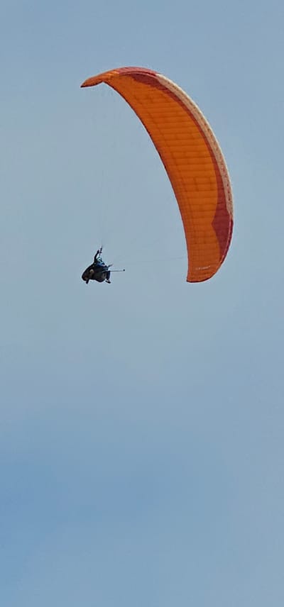 A paraglider soars against a clear blue sky. The paraglider is orange with some red tones, and a person is suspended below it by lines. The location is likely in the vicinity of Alto Hospicio, Chile, considering the region's popularity for paragliding.