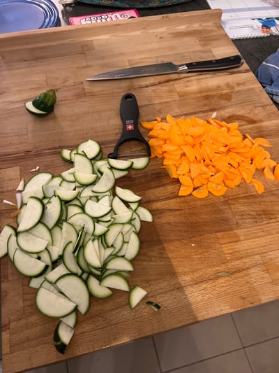 An overhead view captures a wooden cutting board on a kitchen counter, showing ingredients prepared for cooking. A large pile of thinly sliced zucchini or cucumber, cut into semi-circles, occupies the lower right portion of the board. Above and to the left of the zucchini, there is a heap of thinly sliced carrots, also primarily in round or half-round shapes.
A kitchen knife with a black handle lies on the left side of the cutting board, parallel to its edge. A black vegetable peeler, prominently displaying a red Swiss cross logo and the text "SWISS MADE" on its handle, is situated between the piles of carrots and zucchini. A small, green end-piece of a vegetable is visible at the bottom center of the board.
The scene depicts food preparation in progress. In the background, partially visible on the far left, are a blue plastic lid or container and a pink food package, on which the brand "Milka" is clearly legible, with "Oreo" faintly visible above it. A spiral-bound notebook or planner with some text and an image of people is also partially visible at the top left. The indoor lighting suggests a typical kitchen environment. The "SWISS MADE" peeler is a notable detail consistent with the Chêne-Bourg, Switzerland location.