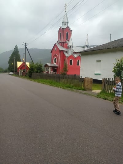 An eye-level, medium shot captures a cloudy day on a paved road in Luhy, Ukraine. The road extends into the distance, bordered by a wooden fence and green grass. On the left, a small red building with a golden roof and cross stands near a larger red church with silver spires and crosses. A white building with a gray roof is visible to the right of the church. A young boy in a striped shirt and beige pants stands on the right side of the road, holding a small object. A white car is parked on the left side of the road. Power lines crisscross the sky. The background shows rolling hills covered in trees.