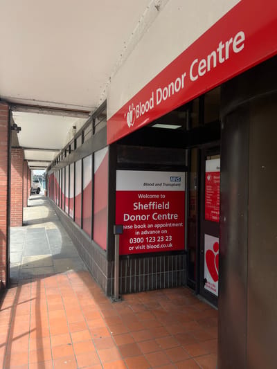 The image shows the entrance to the Sheffield Blood Donor Centre in the United Kingdom. The center is identifiable by the large red and white sign above the entrance, which reads "Blood Donor Centre" in white text on a red background. Further signage near the entrance welcomes visitors to the Sheffield Donor Centre, affiliated with the NHS Blood and Transplant service. Contact information, including a phone number (0300 123 23 23) and website (blood.co.uk), is also displayed on the entrance signage to encourage appointments. There is a shadow cast across the ground, obscuring the terracotta colored tiles. At the end of the alleyway in the distance, a person can be seen walking away from the camera.