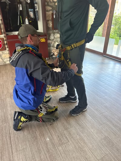 The image shows two people indoors, engaged in the process of wearing climbing harnesses. The person closest to the camera is wearing a grey and blue jacket, a cap, and is adjusting the harness around their waist. Climbing gear is attached to the harness. They have dark-colored mountaineering boots on their feet. The other person is wearing a dark green outfit and black sneakers and is also being fitted with a harness. They are standing closer to a window. The floor is covered with light-colored wood-like tiles. In the background, there appears to be a stone wall and a glass display case containing various outdoor gear. The location is implied to be in Pejë, Kosovo, based on user context.
