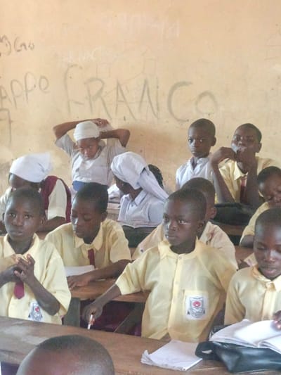 An interior shot depicts a classroom in Mada, Nigeria, featuring approximately ten to twelve individuals, predominantly school-aged children, seated at wooden desks. The room's background consists of a light-colored, worn wall with visible cracks and handwritten graffiti, including the word "FRANCO" prominently. Most children in the foreground wear light yellow, collared short-sleeved shirts, some with maroon ties; one child's shirt displays a school crest on the left pocket. Individuals in the mid-ground and background, likely older students or adults, wear light-colored shirts and white head coverings, with one person in the back adjusting their head covering. Various children are observed looking forward, with one holding a white pen over paper on a desk, and another with an open book and a dark bag on their desk. The lighting suggests an indoor daytime setting.