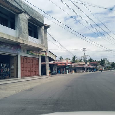This image captures a street scene in a commercial area, likely in Keudealuerheung, Indonesia, under an overcast sky during daytime. The foreground features an asphalt road with minimal traffic.

On the left, a two-story concrete building houses businesses. The ground floor clearly displays a sign for "LIA KOSMETIK," advertising "KOSMETIK DAN SKINCARE," "PERLENGKAPAN MANDI (SABUN & DETERGEN)," and "PERLENGKAPAN BAYI DAN LAIN-LAIN." Contact details, including "Wa: 0822 8061 0887," "Tiktok: @liana.la302," and "Fb: Liana lia," are also visible. Adjacent to the cosmetics shop, a large brown folding door suggests another business entrance, above which a sign for "MIE PANGSIT CABANG REX" is displayed, advertising "Mie Pangsit."

Further down the street on the right, a series of single-story shops or stalls with various roof types line the road. Signs for "WATER ROOM HALIMUN" and a smaller "APEL" sign are discernible, along with a partially visible sign that reads "BS" (possibly for a banking agent). Several motorcycles are parked near these establishments. A few people are visible, including one person riding a motorcycle further down the road and some individuals near the stalls.

Numerous black electrical wires crisscross the frame against the cloudy sky, connected to a power pole in the mid-ground. Palm trees and other tropical foliage are visible in the background, behind the row of shops, reinforcing the Southeast Asian setting. The scene depicts typical daily commerce and local activity.