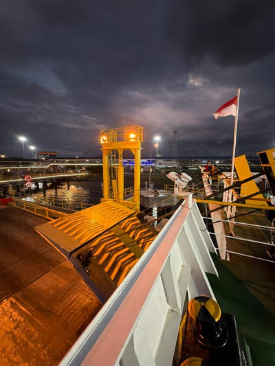 The image depicts a nighttime scene at a port in Melaya, Indonesia, likely a ferry terminal. The sky is overcast and dark, with some streaks of lighter clouds. A red and white Indonesian flag is prominently displayed on a flagpole to the right of the frame, rippling in the wind.
In the foreground, part of a white ship's railing and a black and yellow bollard are visible. Extending from the ship is a large yellow ramp, which appears to be a vehicle ramp for a ferry. This ramp has diagonal black and orange reflective markings. Beyond the ramp, there is a yellow gantry structure with lights.
In the middle ground, the water of the port is visible, reflecting the lights from the shore. A walkway or pier extends into the water, lined with lights. On this pier, a sign with a red circle and a diagonal line with the number "20" inside is visible, indicating a speed limit. Further back, there are buildings and structures associated with the port, including what appears to be a terminal building with the word "PORT" illuminated in blue. Two individuals are standing on a raised platform in the middle distance, possibly port workers.
The overall impression is a functional, active port at night, with a sense of transition and movement. The lighting from the port structures and the ship create dramatic highlights and shadows.