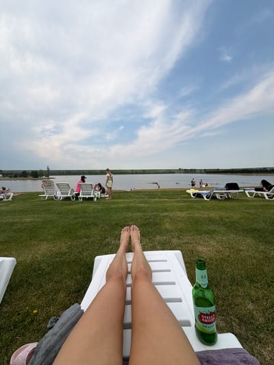In Yemelyanovo, Russia, a person's legs are stretched out on a white lounge chair, facing a lake with people swimming and sunbathing in the background. A green bottle of Stella Artois non-alcoholic beer rests on the chair next to the person's legs. The scene is set on a grassy area adjacent to the lake, under a partly cloudy sky.