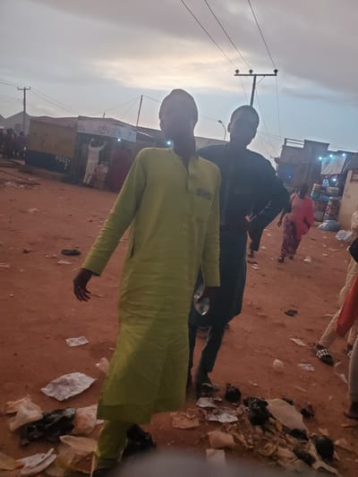 The image depicts an outdoor scene in what appears to be a market area in Funtua, Nigeria. The ground is covered in reddish-brown dirt and scattered with various pieces of litter, including plastic bags and paper. In the foreground, two young men are standing. The man on the left wears a long, light green garment with long sleeves. The man on the right wears a dark colored garment. Behind them, other people are visible, some walking and others standing. In the background, there are market stalls or temporary structures, one of which has the word "POS" visible on it. Overhead, power lines are strung between poles, indicating the presence of electricity infrastructure. The sky is overcast with a pale, diffused light, suggesting it may be early morning or late afternoon. The overall impression is of a busy, somewhat unkempt, commercial environment.
