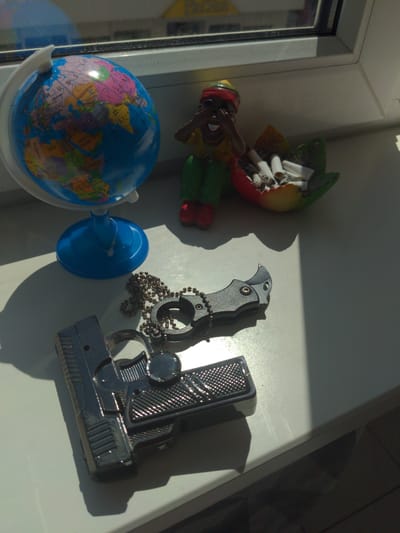 The image shows a collection of objects placed on a white surface, likely a windowsill, in the city of Kamen, Germany. Sunlight casts strong shadows, indicating it is daytime.
In the background, a small globe rests on a blue stand. To its right, a figurine of a person wearing a red headscarf and green pants is positioned next to a colorful, fruit-shaped ashtray filled with cigarette butts.
In the foreground, a metallic toy gun with a chain attached to a small folding knife lies on the surface. The gun has a detailed grip and barrel, and the knife is partially open. The objects are arranged in a static, staged manner.