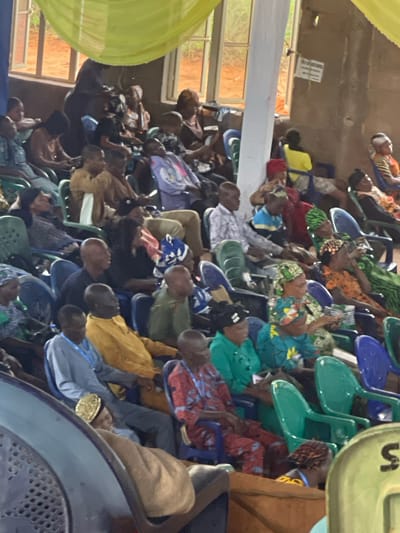 A medium shot, indoors, depicts a crowd of people seated in chairs, seemingly watching an event in Nteje, Nigeria. The people are dressed in a variety of clothing styles. The chairs are mostly plastic, and arranged in rows. Overhead, there are windows to the left with natural light streaming in. To the right, there are balloons and part of a banner visible.