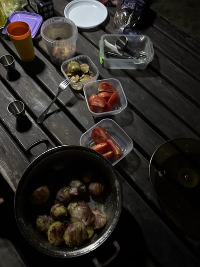 The image depicts a wooden table outdoors, likely at night, with various food items and containers. In the foreground, a pot contains what appear to be potatoes wrapped in bacon. To the right of the pot, a dark, circular object, possibly another pan or bowl, is visible. Further back on the table, there are clear plastic containers filled with sliced tomatoes and what seem to be pickled vegetables. A fork rests on the table near the pickled vegetables. Other items present include a purple plastic lid, an orange and green plastic cup, two small metal shot glasses, and a clear plastic container holding utensils. A white plate is also on the table, along with a bag of bread. The overall scene suggests a casual outdoor meal or picnic. The location is identified as Wołkowiany, Poland.