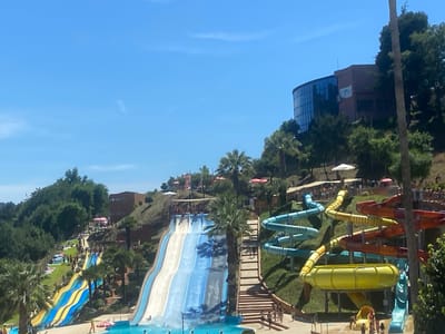 An outdoor water park in Amarante, Portugal is depicted on a sunny day. The image shows several large water slides of various colors and designs, including a multi-lane white and blue slide, a yellow and blue striped slide, and several tubular slides in blue, yellow, orange, and red. People are visible on the slides, in the surrounding pools, and walking on pathways. Lush green trees and vegetation cover the hilly terrain, with some buildings also visible in the background, including a modern building with a curved glass facade. Umbrellas and lounge chairs are scattered throughout the park, indicating areas for relaxation. The sky is clear and blue.