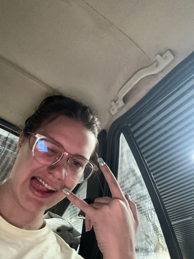 A selfie is taken from inside a vehicle, looking upwards towards the car's ceiling and sunroof. The subject, a young person wearing glasses, is in the foreground, tilted to their right. They have brown hair and are wearing a light-colored t-shirt. Their mouth is open, with their tongue sticking out playfully, and they are winking. One hand is raised, with two fingers extended and pointing towards the camera or the viewer. The person's fingernails are painted a light, shimmery color.
The interior of the car is visible, with the beige headliner and a white grab handle on the left. The sunroof is open, revealing a striped patterned shade overhead and a bright, possibly cloudy, sky. The background outside the sunroof is blurred but appears to be some sort of structure with vertical elements. The lighting suggests it is daytime. The overall mood is lighthearted and energetic. No visible text is present in the image.