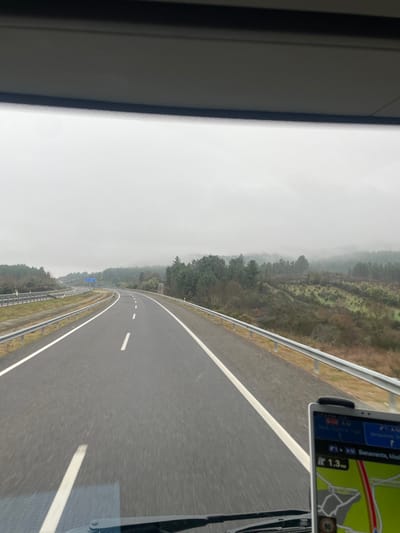 The image depicts a view from the interior of a vehicle traveling eastbound on a multi-lane highway, likely the A-24 in the vicinity of Chaves, Portugal, heading towards the Spanish border. The GPS device visible on the lower right of the windshield indicates a route towards "A-52 Benavente, Madri" and "Vigo A-52," with a distance of "1.3 mi" to an unspecified point on the route.
The scene is dominated by a two-lane asphalt highway featuring white solid edge lines and white dashed lane dividers, suggesting a single direction of travel. Metal guardrails line both sides of the roadway. A blue highway sign is partially visible in the distance on the left side of the road. The vehicle's windshield, dashboard, and a portion of the interior roof lining are visible in the foreground. No individuals are discernible.
The environment is characterized by a rural, hilly landscape under heavy overcast or foggy conditions, resulting in a uniformly gray sky. The hills flanking the highway are covered with a mix of evergreen trees and bare deciduous vegetation, some displaying reddish-brown tones. The distant terrain is obscured by mist or low clouds. The overall impression is one of movement along a highway through a forested, undulating landscape on a dim, possibly damp, day.