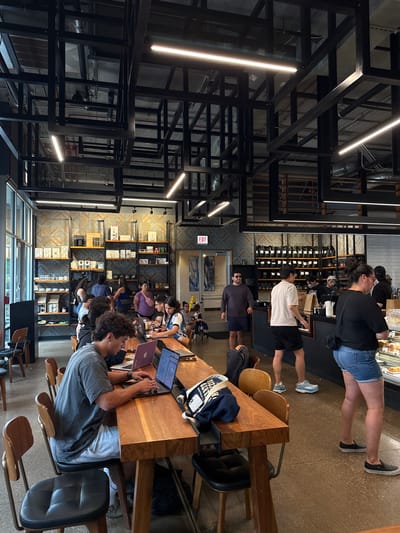 The image captures the interior of a busy coffee shop, likely in Naperville, United States of America, during the daytime. The architecture features an industrial design with exposed black metal beams and ductwork supporting the ceiling, interspersed with modern linear light fixtures. The walls are adorned with geometric patterned wallpaper, and shelves displaying coffee-related products and packaged goods line the back wall.

In the foreground, a long, wooden communal table dominates the space, with several patrons seated and engaged in various activities. A young man in a grey t-shirt and light grey shorts is seated at the table, focused on his laptop, which is open. Next to him, another individual is also working on a laptop. The table is set up for multiple users, with chairs spaced along its length.

Behind the table, a counter area is visible where staff are serving customers and preparing orders. Several people are standing at the counter, including a man in a white t-shirt and black shorts, and a woman in a black t-shirt and denim shorts, who appears to be looking at a display case. To the right, more patrons are standing, including a woman in a black top and blue jeans.

The environment is well-lit, with natural light streaming in from large windows on the left side of the image. The overall atmosphere suggests a lively, contemporary coffee shop environment where people gather to work, socialize, and enjoy beverages. The presence of multiple laptops and the general activity indicate a typical urban coffee shop setting.