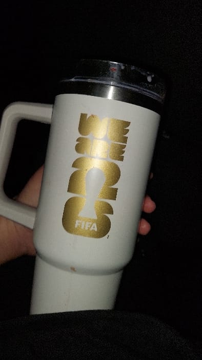 A close-up image depicts a white, insulated travel mug with a light grey handle, held by a human hand. The mug features a prominent gold-colored logo on its side, which reads "WE ARE ONE" with the "O" or "0" stylized as a FIFA World Cup trophy silhouette, followed by "FIFA" in smaller text below. The mug's lid is silver-colored with a clear plastic top, exhibiting visible splashes or residue of a white, milky liquid. The background is uniformly dark and indistinct, suggesting an interior environment with low light conditions or a deep focus on the object. A dark, possibly fabric surface, is visible in the lower foreground. No specific location within Tlaxcala City, Mexico, is discernible from the visual evidence provided.