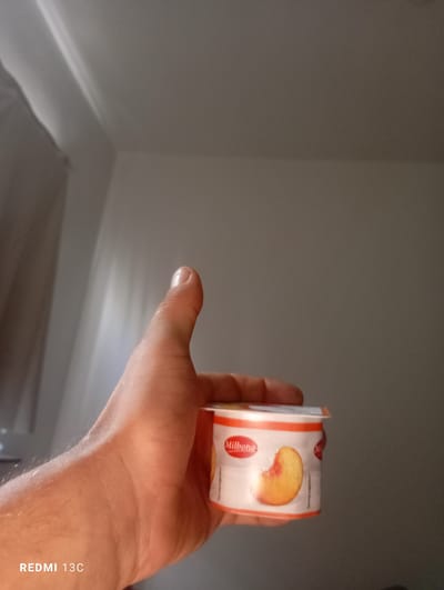 A hand holds a small, single-serving yogurt cup featuring a picture of a sliced peach on its label. The yogurt cup is white with orange accents and has the brand name "Milbona" in red text. The hand is a fair-skinned adult hand, with visible hair on the forearm. The background is a dimly lit, neutral-colored wall, possibly an interior room. Light is streaming in from an unseen source, creating bright highlights on the wall and casting subtle shadows. The "REDMI 13C" text at the bottom left indicates the device used to take the photo. The overall impression is a simple, candid shot of a person holding a snack.