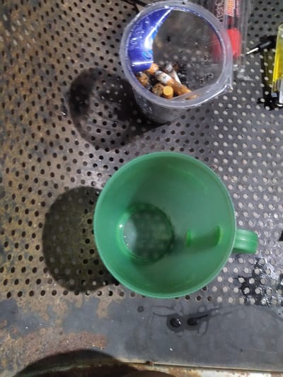 An overhead, medium shot shows a clear plastic cup filled with cigarette butts on a perforated metal surface. To the right of the cup, a red marker and a yellow object are visible. In the lower right quadrant, a green plastic cup is centered on the surface, casting a shadow. The metal surface appears rusted in some areas and wet in others, with shadows of the cups cast on it. The overall lighting is dim. The image is taken from an elevated perspective, looking down on the scene. This scene is likely taking place in Gampongbaro, Indonesia, where such a collection of discarded items could be found in an outdoor or workshop setting.