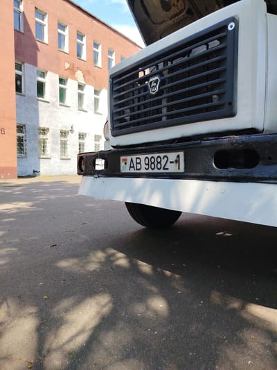 A low-angle, close-up shot captures the rear of a white truck parked on an asphalt surface, likely in the city of Biaroza, Belarus. The truck's black bumper is visible, adorned with a white license plate that reads "BY AB 9882-1." The grill of the truck, featuring the GAZ logo, is also in frame. In the background, a two-tone building with salmon-pink and white walls stands, featuring several windows with bars. The sky above is a pale blue with scattered clouds. The asphalt is marked by shadows cast by the truck and overhead foliage, with small debris scattered across its surface.