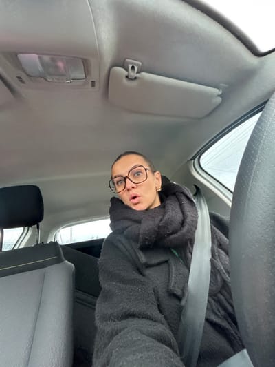 A female individual is depicted seated in the front passenger seat of a vehicle in Saint-Cyprien, France. She is wearing dark, tortoise-shell rimmed eyeglasses and a thick black coat and scarf. Her dark hair is pulled back, and a gold-toned earring is visible on her right ear. Her mouth is open in an 'O' shape, and she looks directly at the camera. A light grey seatbelt is secured across her chest and shoulder. The vehicle's interior features a grey headliner, an overhead light unit, and a folded grey sun visor on the passenger side. The upper portion and headrest of the driver's seat, upholstered in grey fabric with yellow-green stitching, are visible to the left. The rear window reveals an overcast exterior with visible water droplets on the glass. A dark, possibly upholstered, parcel shelf or rear seatback is visible in the background.