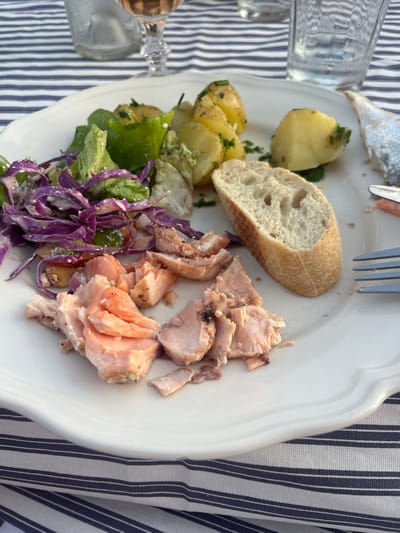 A forensic description of the scene reveals a meal set on a table, likely in Skagen, Denmark, as indicated. A white, scalloped-edge plate occupies the center, laden with various food items. On the left side of the plate, flaked, cooked salmon, appearing light pink and orange, is distributed in irregular pieces. Adjacent to the salmon is a portion of salad, comprising green lettuce leaves and shredded purple cabbage, possibly coated in a light dressing. To the rear of the salad, several small, whole or halved potatoes, yellowish in color and sprinkled with green herbs (likely chives or parsley), are present. A single piece of a white baguette, approximately 10-15 cm long and cut lengthwise, rests on the plate to the right of the potatoes and in front of a metal fork with four tines. A partial view of what appears to be the head or skeletal remains of a smaller fish is visible on the far right edge of the plate, suggesting another component of the meal. The plate rests on a blue and white striped fabric tablecloth. In the blurred background, two clear glasses are visible: one appears to contain water, and a stemmed glass holds a rosé-colored liquid, likely wine. The arrangement indicates a meal in progress or recently concluded.