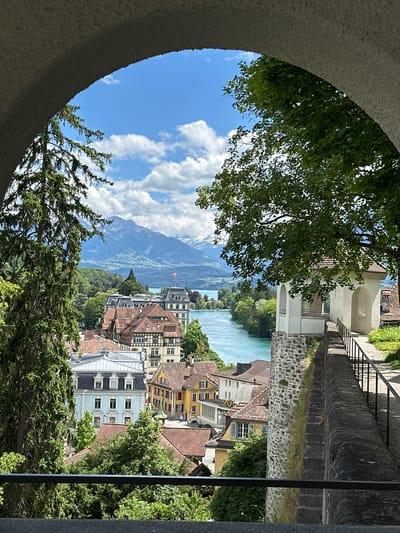 The image depicts a scenic view of the city of Thun, Switzerland, framed by an arched stone structure. In the foreground, the rough texture of the stone archway is visible, with a large evergreen tree on the left and lush green foliage on the right. Beyond the arch, a panoramic vista unfolds. A bright turquoise river, likely the Aare, flows through the city, reflecting the clear blue sky dotted with white, puffy clouds.

The cityscape features a collection of buildings with traditional European architecture, characterized by red-tiled roofs and a mix of architectural styles. Prominent among them is a larger building with a distinctive flag atop its roof. In the distance, a range of majestic mountains, some capped with snow, forms a breathtaking backdrop. The scene is serene and picturesque, suggesting a tranquil moment in the Swiss Alps region.

The perspective suggests the photograph was taken from an elevated position, possibly a vantage point overlooking the city and the river. The lighting indicates a bright, sunny day, enhancing the vibrant colors of the landscape. No people are clearly visible in the image, adding to the sense of peaceful solitude.