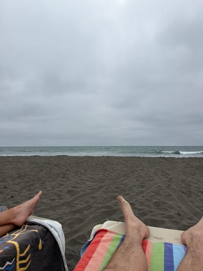 A beach scene unfolds with dark sand meeting the ocean under a gray, cloudy sky. In the foreground, parts of two people are visible, each on a towel. One person is on a striped towel with their feet visible. The other has a towel with a geometric design and the bare feet can be seen. The perspective is from above, looking down towards the ocean.
