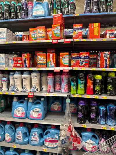 This image captures a retail store aisle, primarily displaying laundry care products. The scene is dominated by shelves stacked with boxes of dryer sheets and bottles of liquid fabric softener or scent boosters. The image is oriented vertically, suggesting it was originally taken in portrait mode and then rotated.

On the left side, shelves are filled with various sizes and types of Bounce dryer sheets, including "MEGASHEETS" and "Tumble" brand sheets. Some boxes have visible price tags and "Rollback" signs indicating sales, with prices like "$3.24" and "$6.97". A sign on the leftmost shelf advises, "Ask for assistance with items on the top shelf."

The right side features an extensive selection of liquid fabric softeners and in-wash scent boosters. Brands visible include Downy, Unstopables, and Fusions 2:1, available in various colors and scents such as "SOFT," "Light Linen," and "Gentle." These bottles are arranged neatly on multiple shelves. Several "Rollback" tags are also present on these items, with prices like "$15.97" visible.

Hanging near the center-right, above some Downy products, are bundles of red plastic cups and packages of "DRYER BALLS," suggesting an adjacent section or cross-merchandising. The overall setting is brightly lit, typical of an indoor retail environment, likely a supermarket or large department store. The shelves are well-stocked, and the products are organized by brand and type.