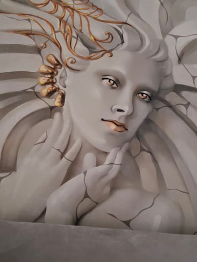 The media file displays a detailed close-up of an artwork, likely a mural or bas-relief sculpture, featuring a stylized ethereal or angelic figure. The central subject is a serene, contemplative face, tilted upwards and to the right, with pronounced, deep-set eyes that gaze slightly upwards. The figure's lips are a subtle peachy-pink, providing a warm contrast to the pale, alabaster-like skin tone.
Elaborate golden, leaf-like or feathery adornments frame the figure's head, sweeping upwards and to the right, resembling either intricate hair or a divine headdress. Matching golden accents are visible on what appear to be fingernails on hands that gently frame the figure's head and neck. The figure and its integrated background are rendered in soft, muted tones of off-white, light grey, and beige. Intricate dark lines, resembling cracks or veins, traverse the figure's skin, hands, and the swirling, organic forms of the background, evoking the appearance of aged marble, shattered porcelain, or ancient stone.
The artwork exudes a dreamlike quality, blending classical sculptural elements with fantasy aesthetics. The rendering creates a strong sense of three-dimensional depth and form. The lighting emphasizes the contours of the face and the metallic luster of the golden details. No visible text is present in the image. The artwork appears to be part of an indoor installation, located in Waldmünchen, Germany.