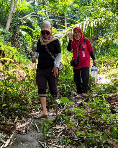 Two women, both wearing hijabs, are walking on a muddy path in a dense forest in Gunung Ambat, Indonesia. The woman on the left, wearing sunglasses and a black t-shirt with a light grey long-sleeved shirt underneath, is walking ahead. The woman on the right, wearing a bright pink t-shirt and a red hijab, is waving at the camera and carrying a clear plastic water jug in her right hand. Both women are wearing dark-colored pants and sandals. The path is surrounded by lush green vegetation, with sunlight filtering through the leaves of the trees, creating a dappled effect. Fallen leaves and bamboo stalks litter the ground. The scene appears to be a casual walk or hike in a natural environment.