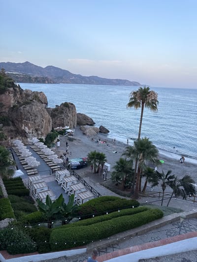 The image depicts a scenic view of a beach resort in Nerja, Spain. The scene overlooks a sandy beach with scattered rocks, leading to the clear blue ocean. Several palm trees are dotted along the shoreline, adding to the tropical ambiance. In the foreground, a neatly arranged row of white lounge chairs with umbrellas suggests a relaxing beach setting. Beyond the chairs, manicured green hedges and decorative outdoor lighting create a sense of order and aesthetic appeal.

The beach itself is populated with a few individuals enjoying the seaside, some wading in the water, while others are seen lounging on the sand or near some beachfront structures. A small blue boat is visible on the sand. In the background, rugged mountains rise up from the coastline, under a pale, hazy sky. The light suggests it is either early morning or late afternoon, with a gentle, diffused light. There is no visible text in the image. The overall impression is one of a tranquil and picturesque holiday destination.
