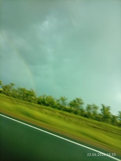 The image captures a diagonal view of a road with a white line, flanked by a blurred green grassy embankment on the left. Beyond the embankment, a row of out-of-focus green trees is visible against a hazy, greenish-blue sky. A faint rainbow arc is present in the upper left portion of the sky. The bottom right corner of the image displays a timestamp: "22.06.2026 19:30". The scene appears to be a roadside in a rural or semi-rural setting, possibly on the outskirts of Sarytogay, Kazakhstan, given the context. The lighting suggests it is late afternoon or early evening, with overcast conditions possibly due to recent rain, indicated by the presence of the rainbow. There are no people or vehicles visible in the frame.