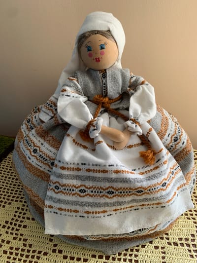 This media file shows a close-up of a traditional Belarusian rag doll, likely homemade, lying on its side. The doll has a simple wooden head with painted blue eyes, rosy cheeks, and a small pink mouth and flower detail. It is dressed in layers of patterned fabric, featuring stripes of grey, white, and rust orange, with some decorative stitching. A white fabric forms a bonnet or headdress around its head and wraps around its body, tied with a brown cord and a small fluffy orange tassel. A small wooden limb, possibly an arm or leg, is visible peeking out from the fabric. The doll is resting on a textured yellow crocheted mat. The background is a plain, light beige wall. The lighting suggests an indoor setting, possibly during the daytime, with no indication of specific weather conditions. The overall impression is one of rustic craftsmanship and cultural heritage. There is no visible text in the image.