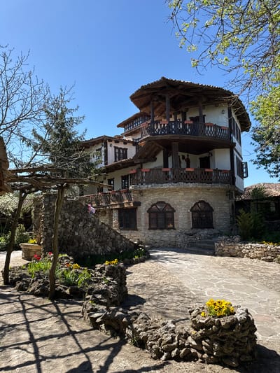 The image captures a three-story building with a distinctive architectural style in Kalimantsi, Bulgaria. The building has a stone facade on the ground floor with arched windows, while the upper floors feature white walls and wooden balconies with intricate railings. A tiled roof tops the structure. In front of the building, there is a stone pathway with flower arrangements in rocky planters. A wooden pergola-like structure and bare trees are visible near the pathway, partially obscuring the building. The sky is clear and blue.