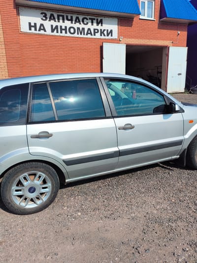 A silver Ford Fusion is parked on a gravel lot in front of a brick building. The building has a blue roof and a sign that reads "ЗАПЧАСТИ НА ИНОМАРКИ" which translates to "SPARE PARTS FOR FOREIGN CARS". The building has two large white garage doors, one of which is open, revealing the interior of a workshop. The car is facing to the right, and its front end is not fully visible. The sun is shining brightly, reflecting off the car's windows. The environment appears to be a commercial area, possibly an auto repair shop.
